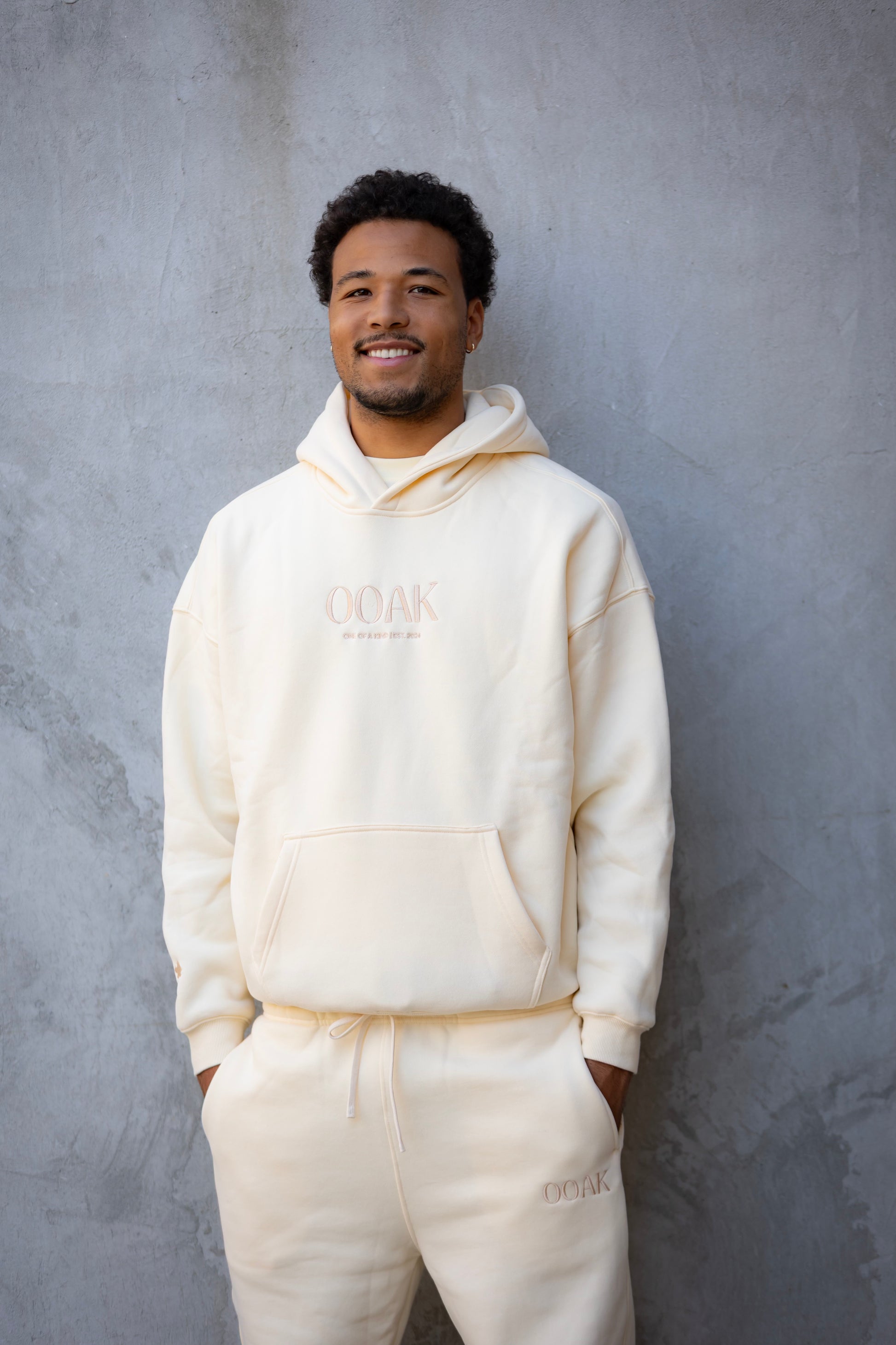 Man wearing a cream-colored hoodie and pants with 'OAK' branding against a gray background