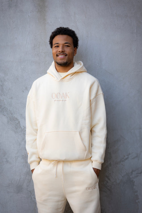Man wearing a cream-colored hoodie and pants with 'OAK' branding against a gray background