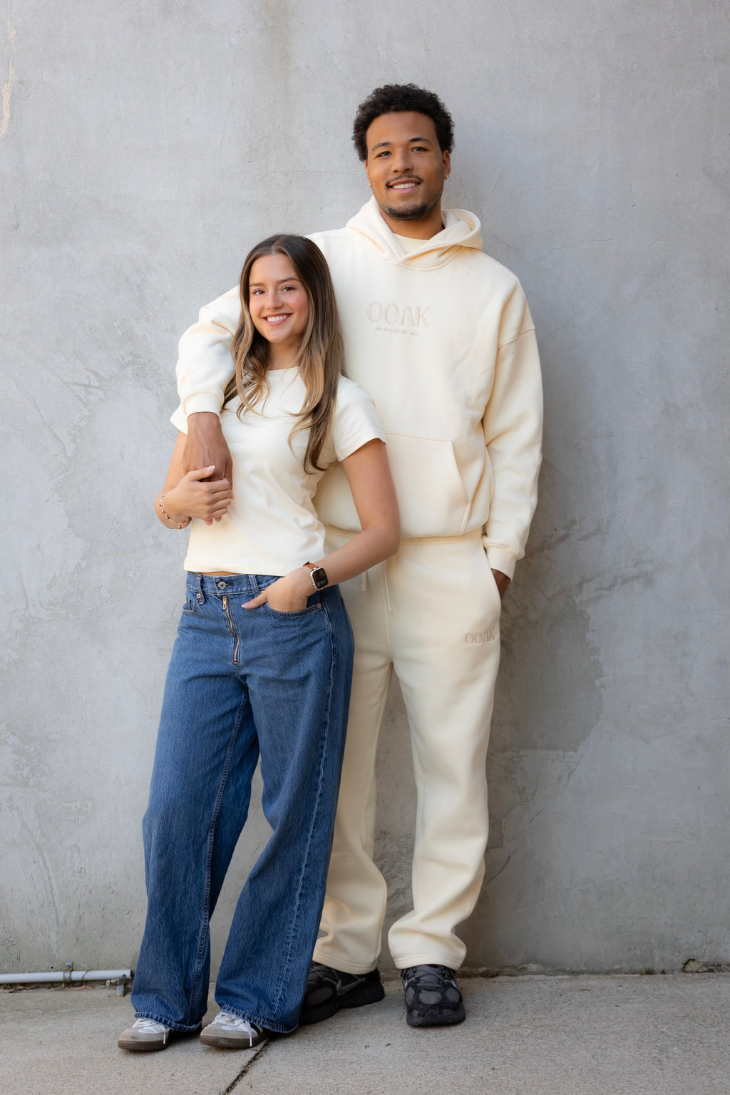Two people standing against a gray wall, one in a white outfit and the other in casual clothing.