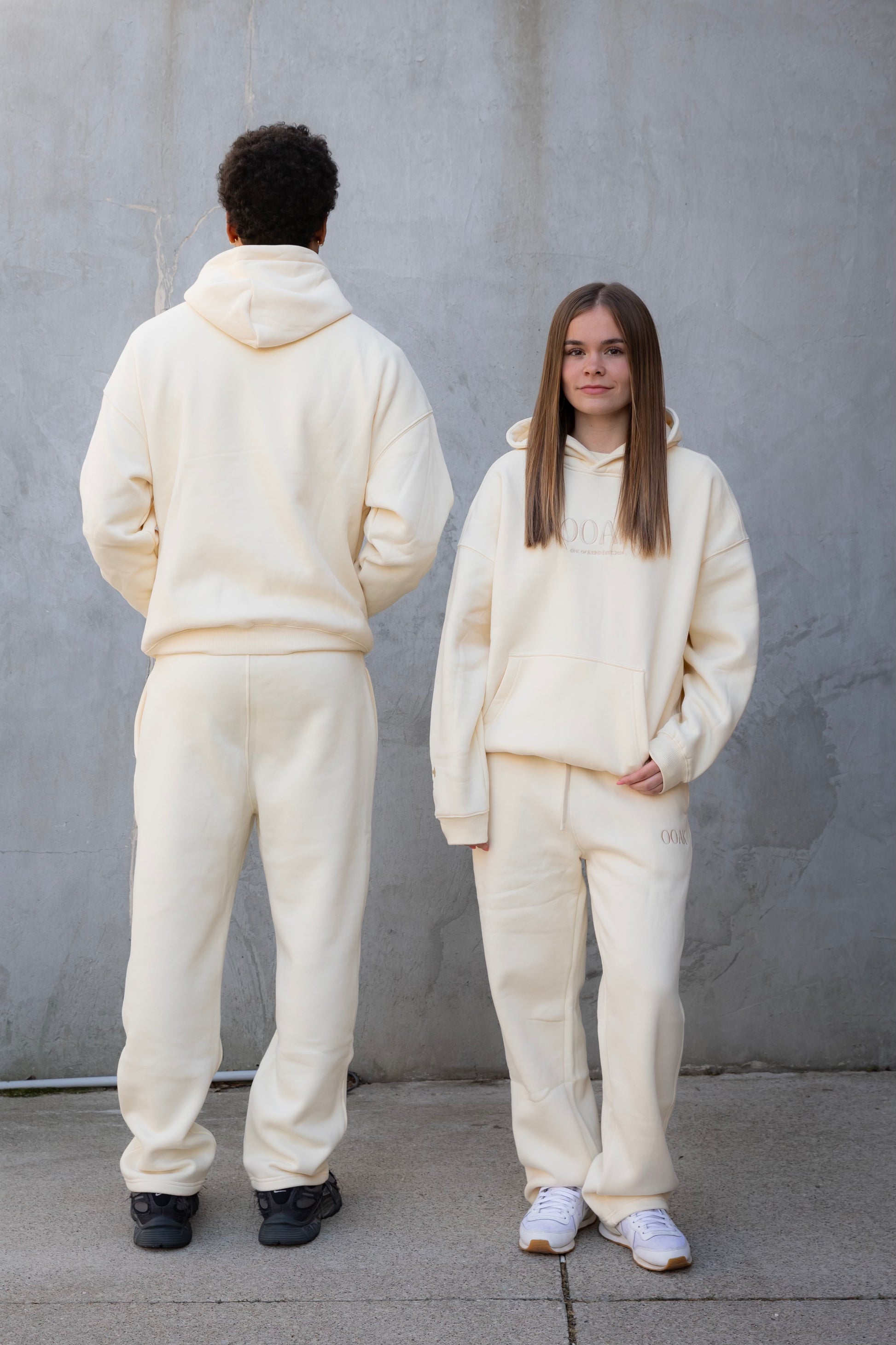 Two people wearing matching cream-colored outfits standing against a gray wall.