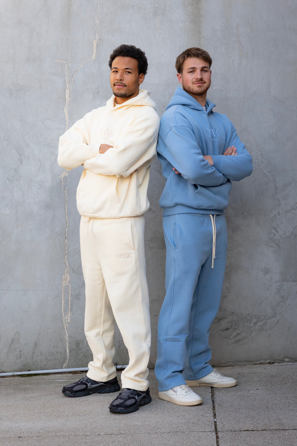 Two men standing back-to-back wearing white and blue tracksuits against a gray wall.