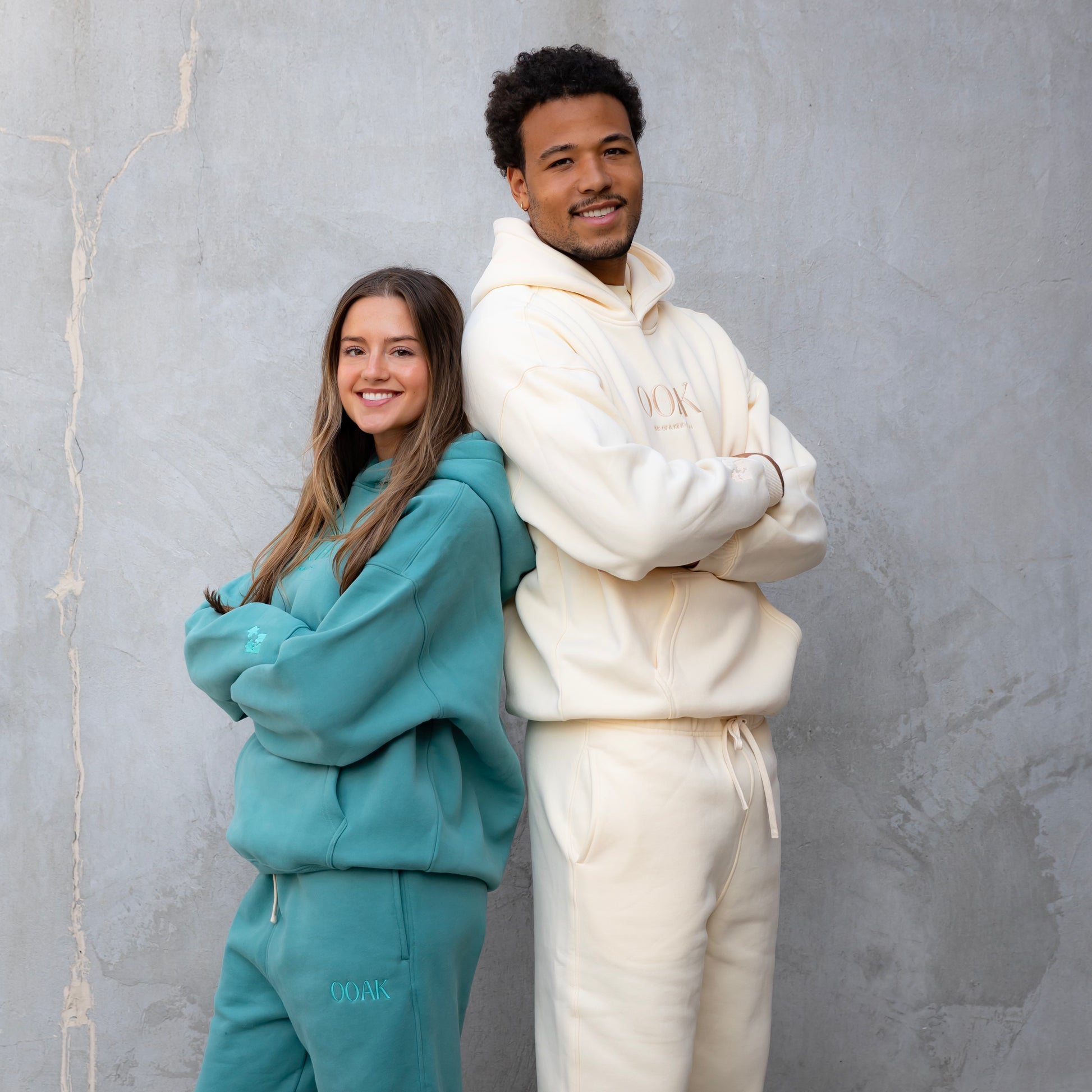 Two people wearing matching teal and white hoodie and pants outfits against a gray wall.