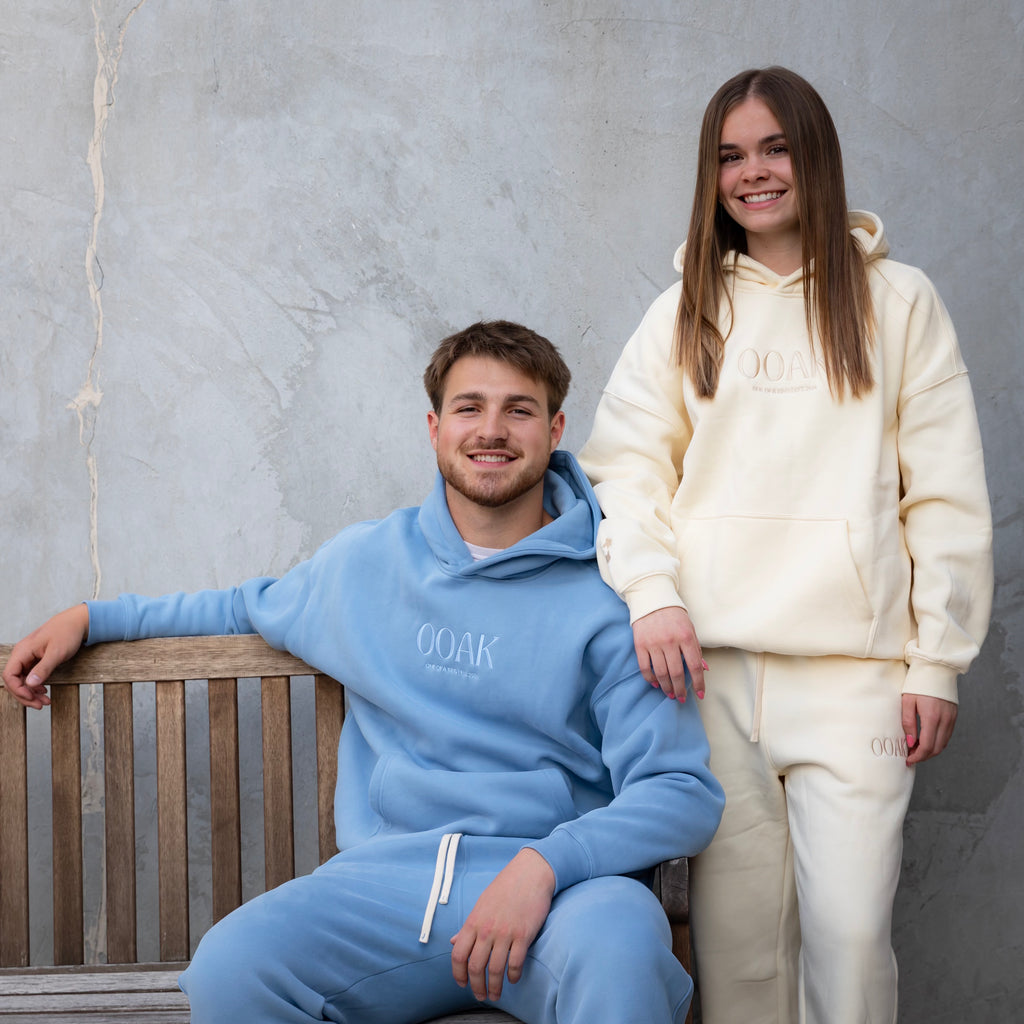 Two people wearing matching blue and white hoodies and pants sitting on a wooden bench against a gray wall.