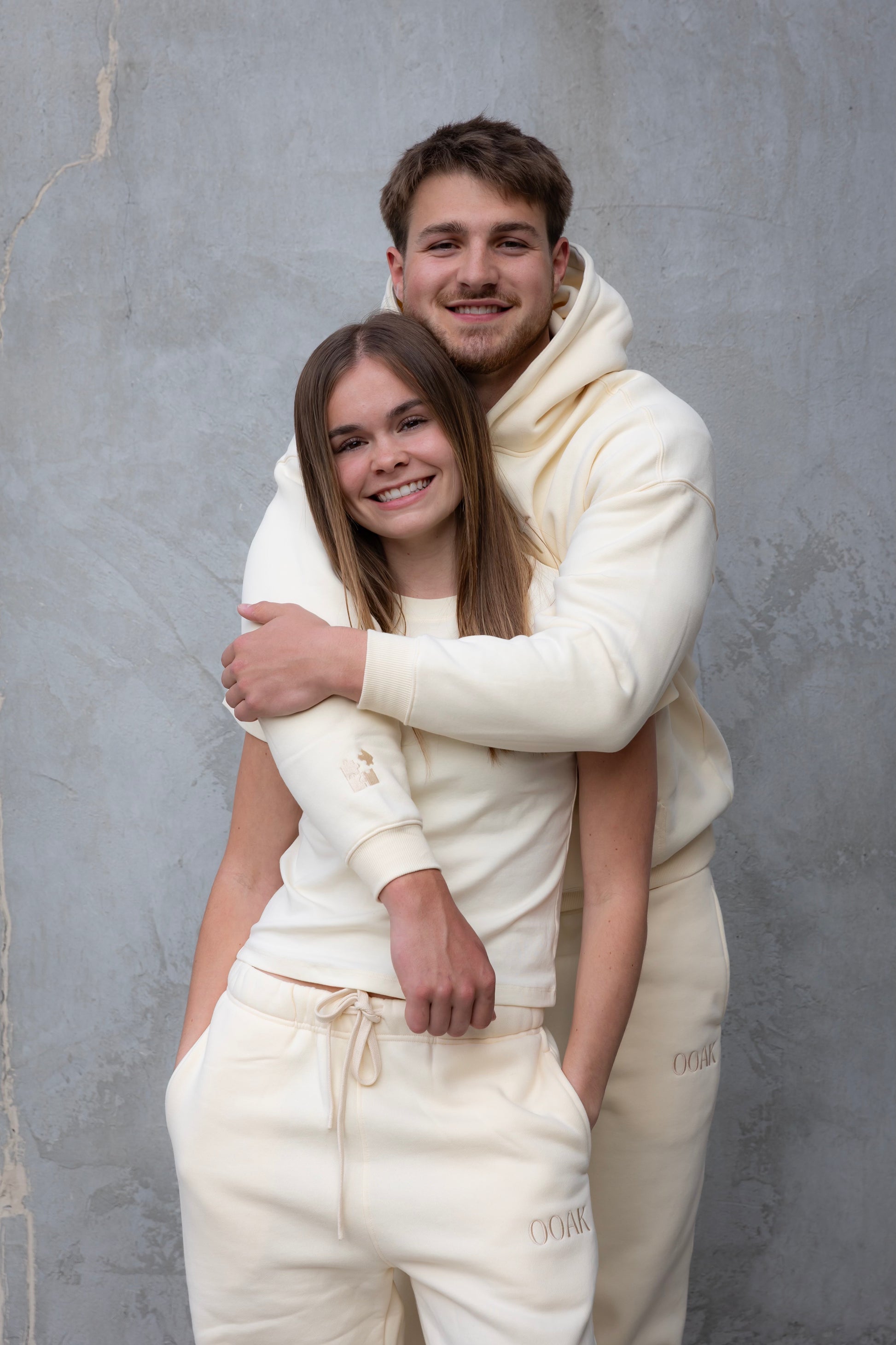 Two people in matching white outfits standing against a gray textured wall.