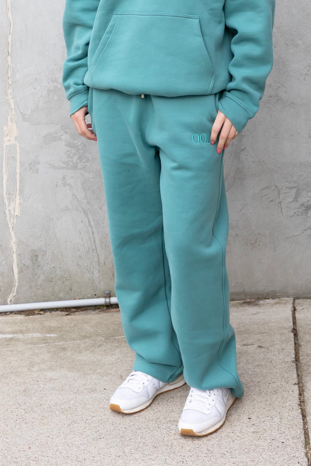 Person wearing a teal tracksuit with white shoes against a gray wall.
