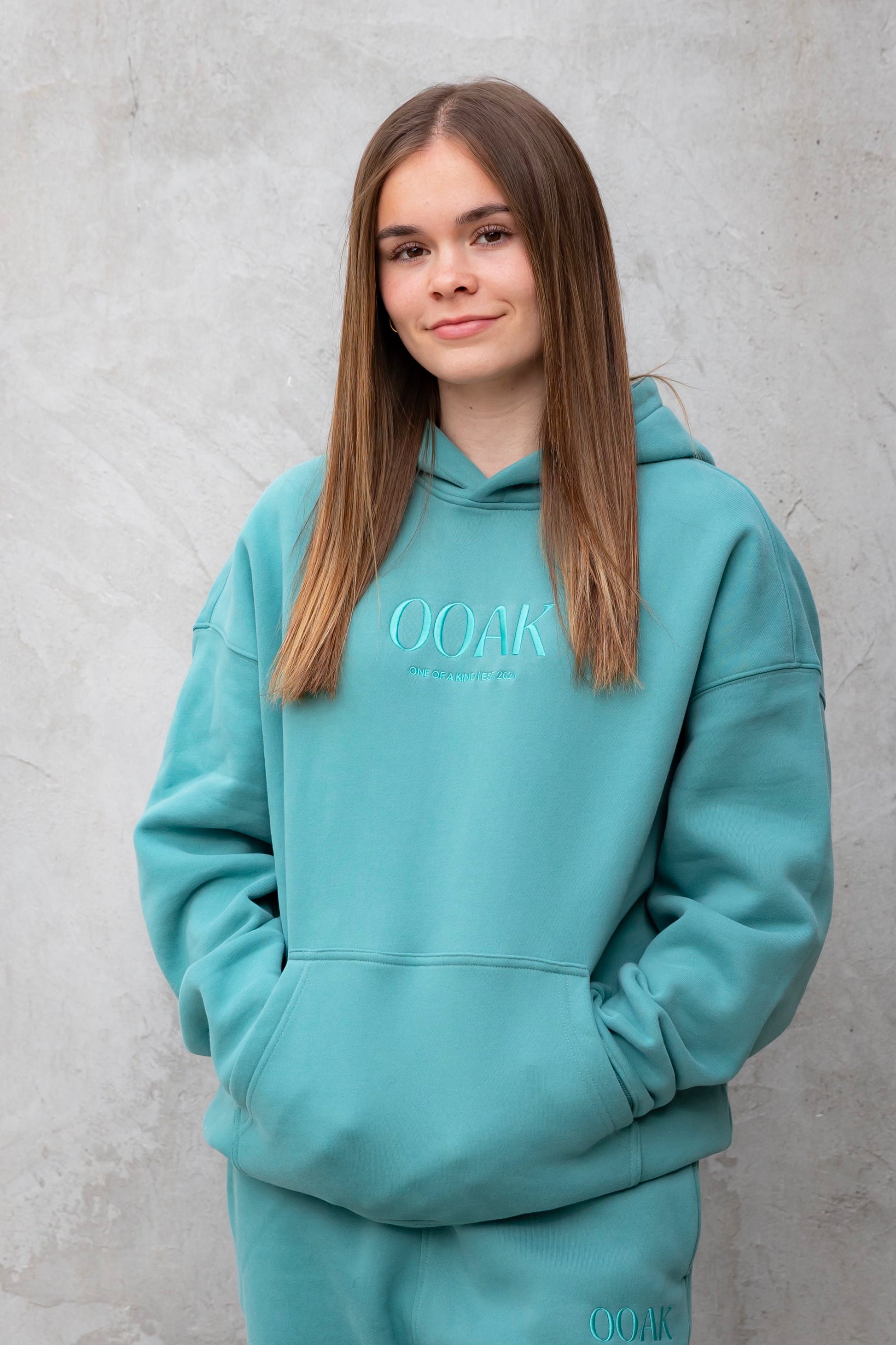 Person wearing a teal hoodie with 'OOAK' branding against a gray background