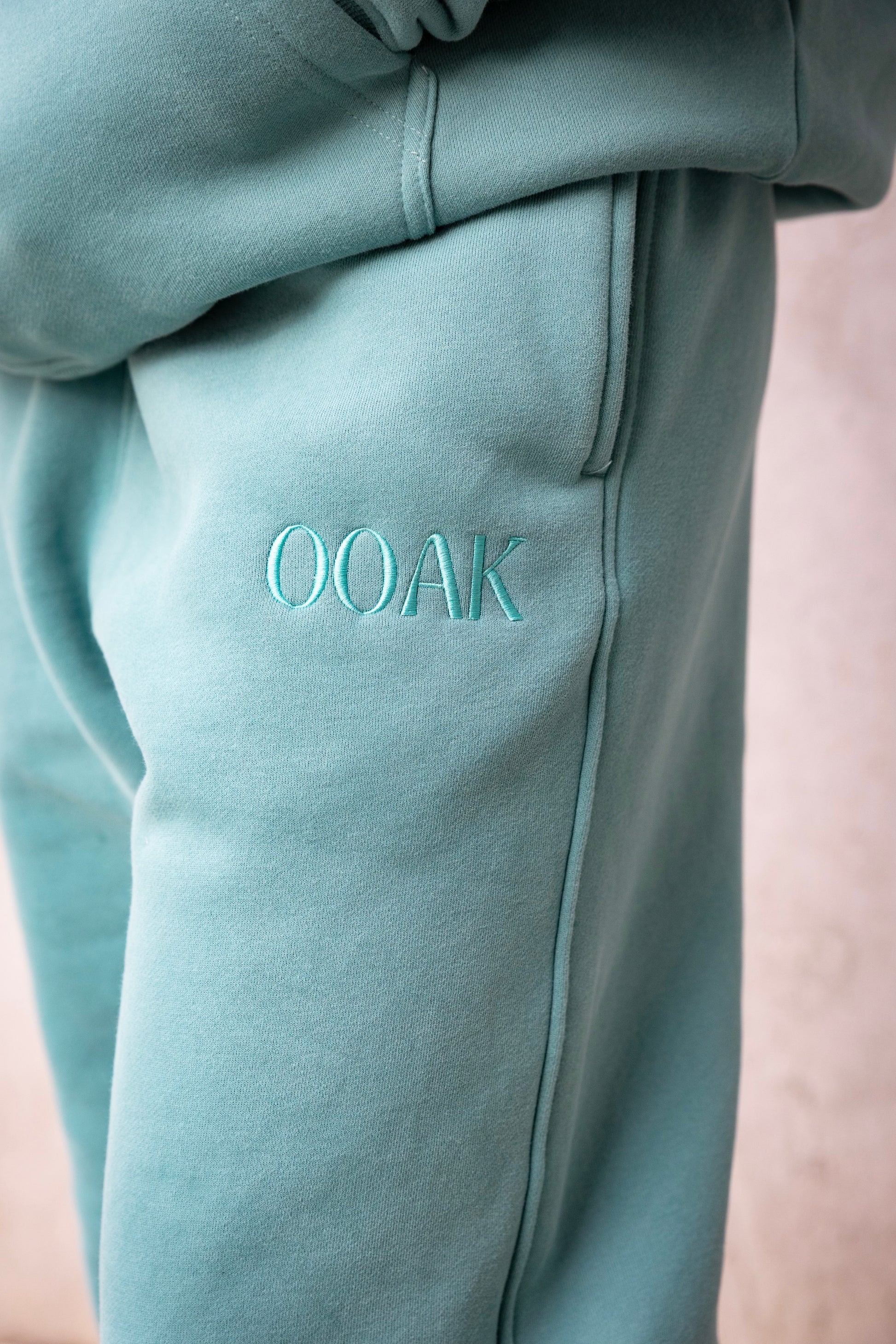 Teal sweatpants with 'OOAK' embroidery on a light pink background