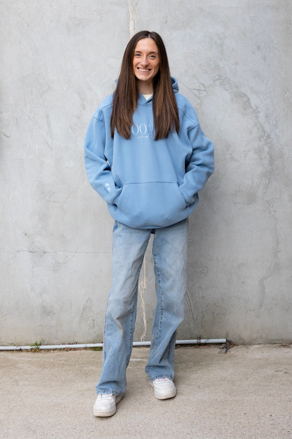 Person wearing a blue hoodie and jeans standing against a gray wall.