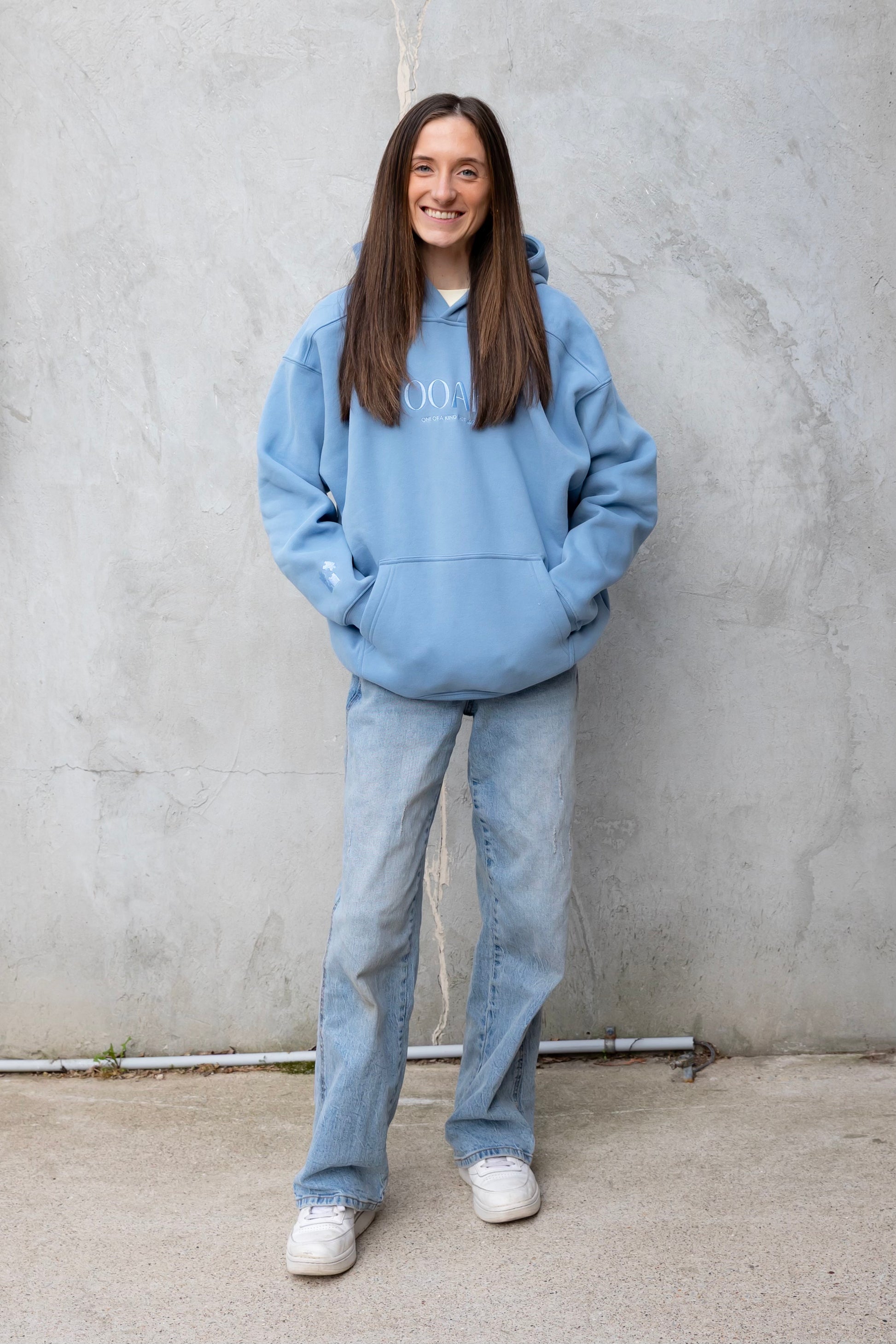 Person wearing a blue hoodie and jeans standing against a gray wall.
