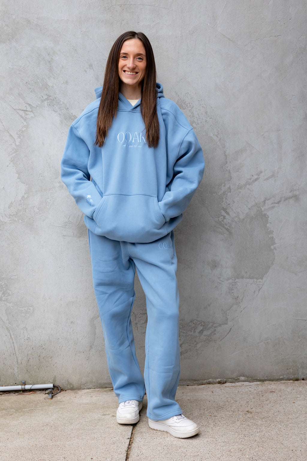 Person wearing a blue hoodie and pants standing against a gray wall.