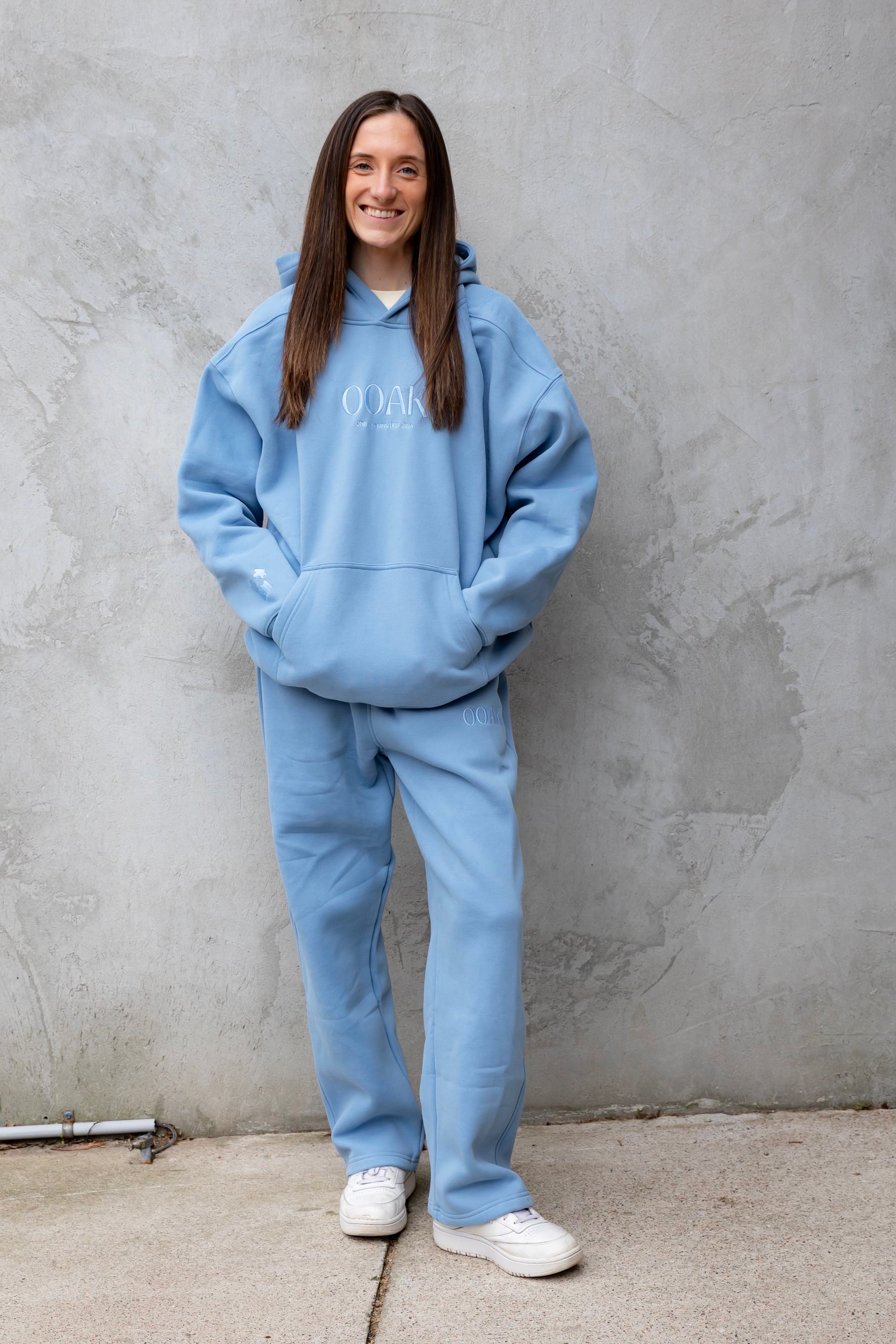 Person wearing a blue hoodie and pants standing against a gray wall.