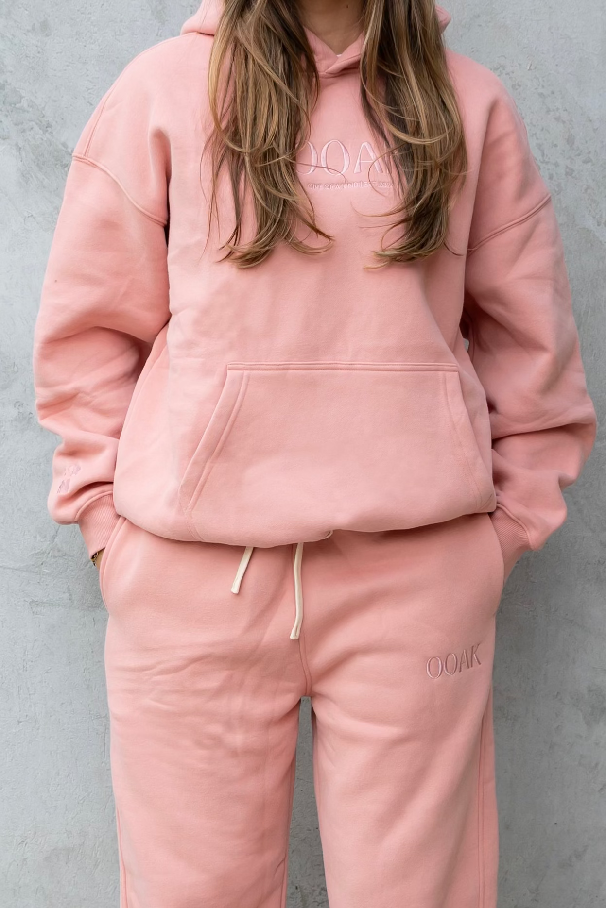 Person wearing a pink tracksuit standing against a gray wall.