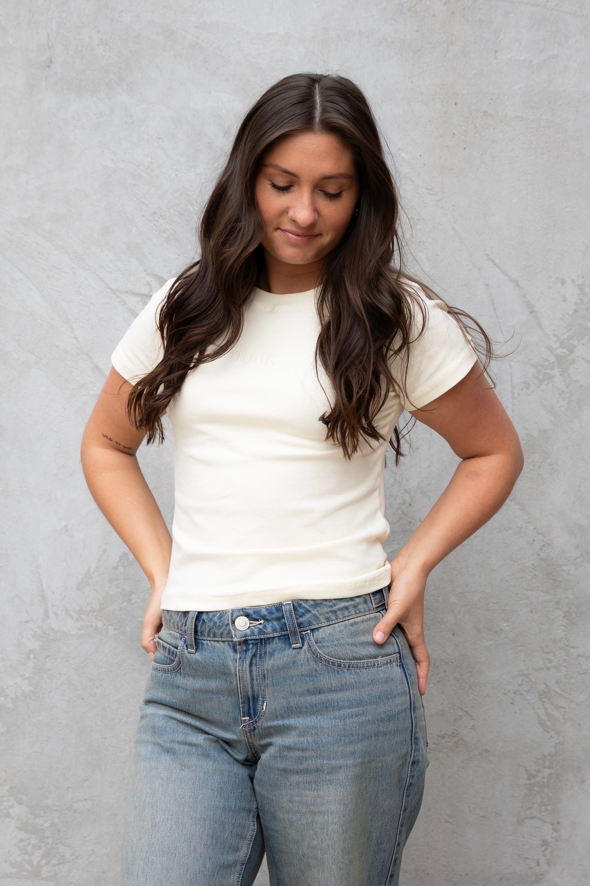 Woman wearing a white t-shirt and blue jeans against a gray concrete wall.