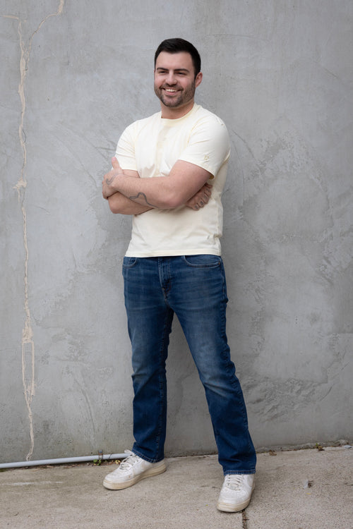 Man wearing a white t-shirt and blue jeans standing against a gray wall.