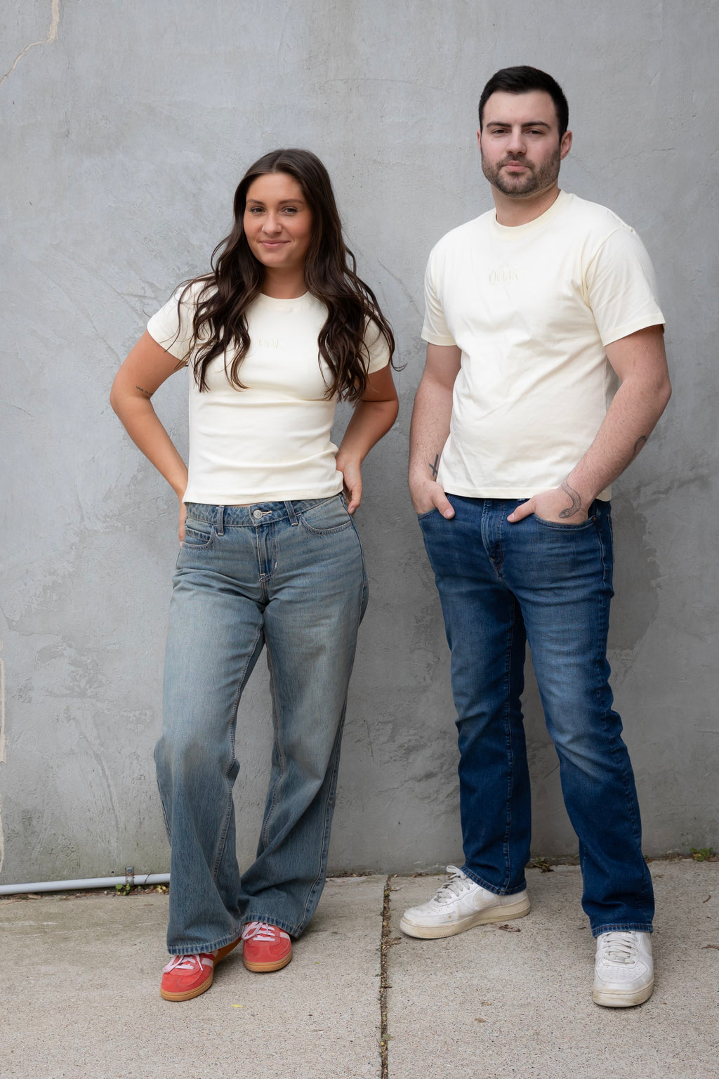 Two people wearing white t-shirts and blue jeans standing against a gray wall.