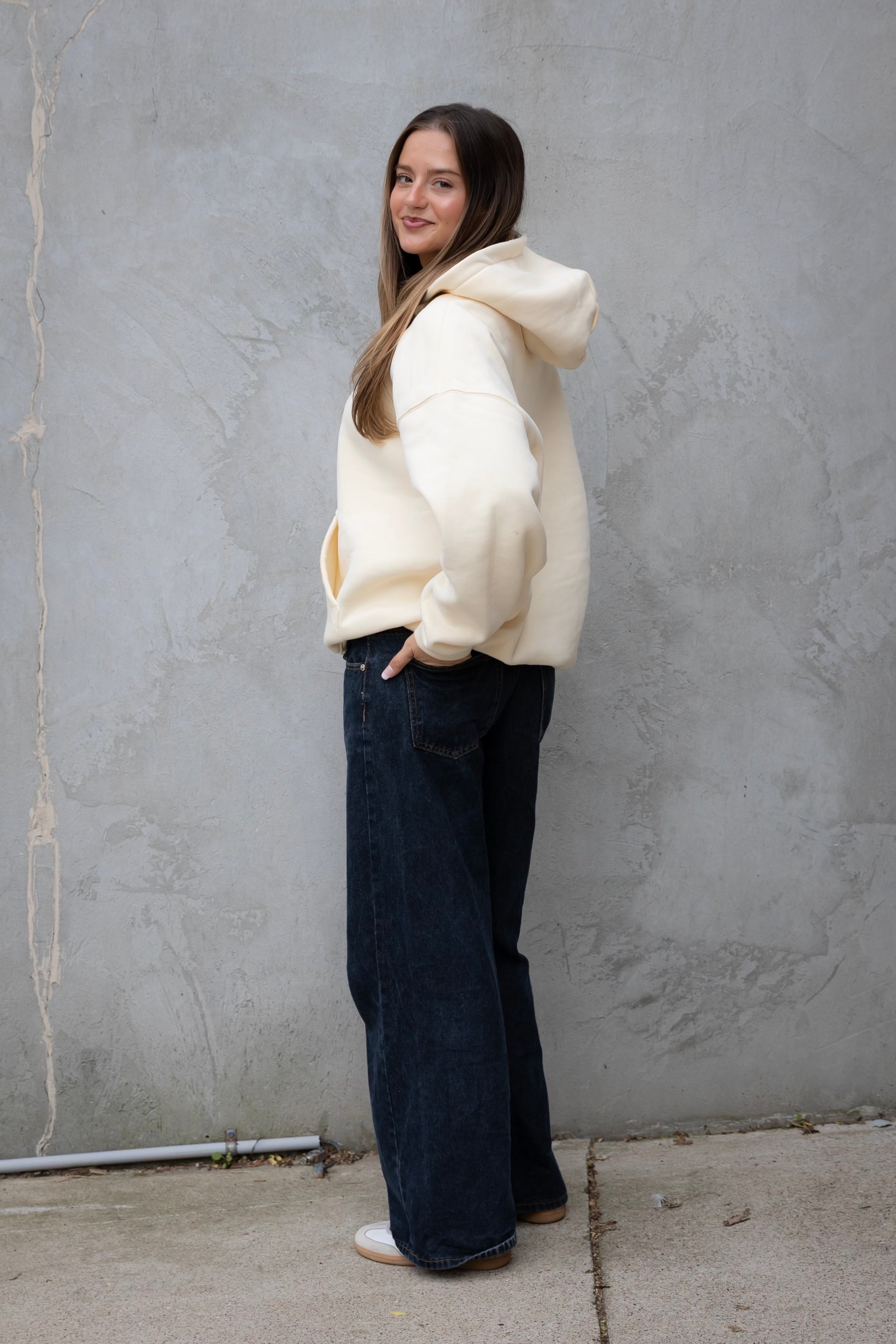 Woman wearing a white hoodie and dark jeans standing against a gray wall.