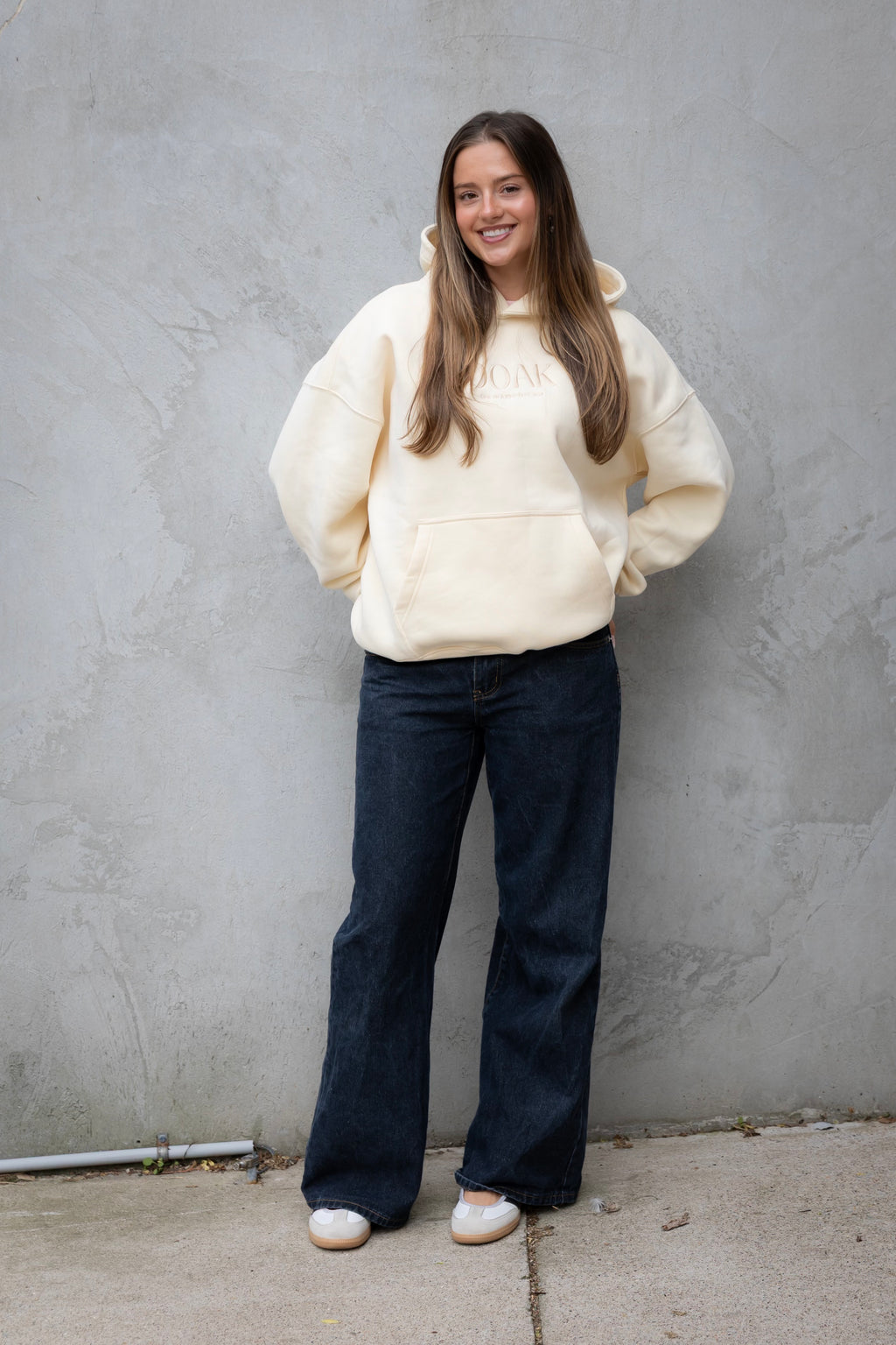 Woman wearing a cream hoodie and dark jeans standing against a gray wall.