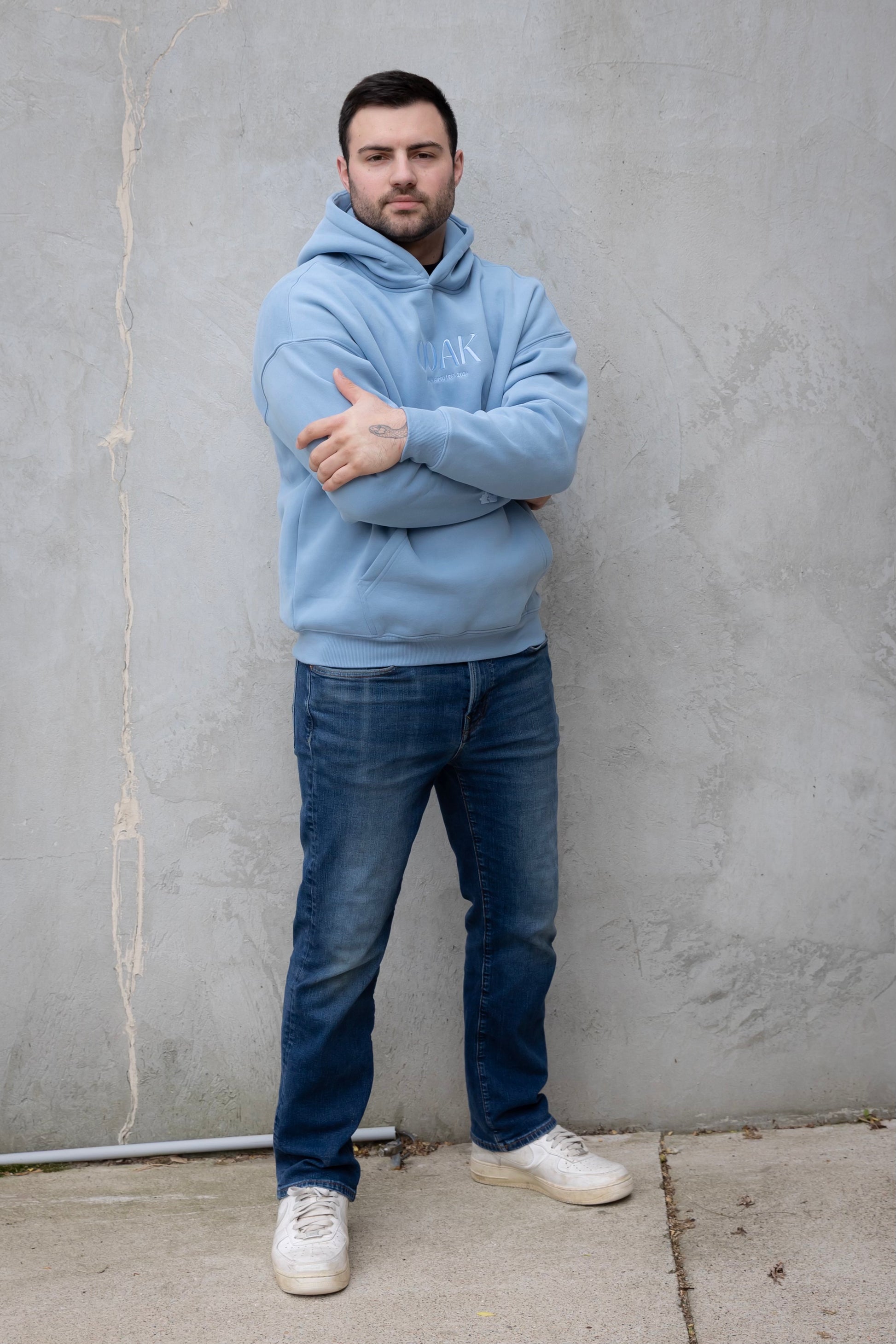 Man wearing a light blue hoodie and jeans standing against a gray wall.