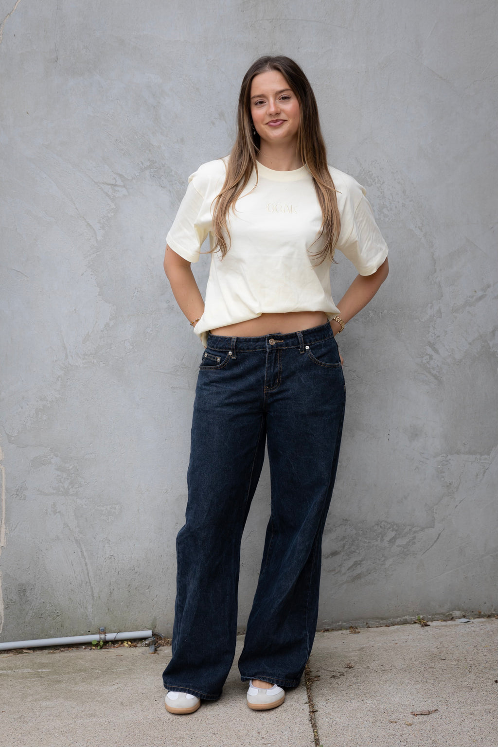 Woman wearing a white top and blue jeans standing against a gray wall.