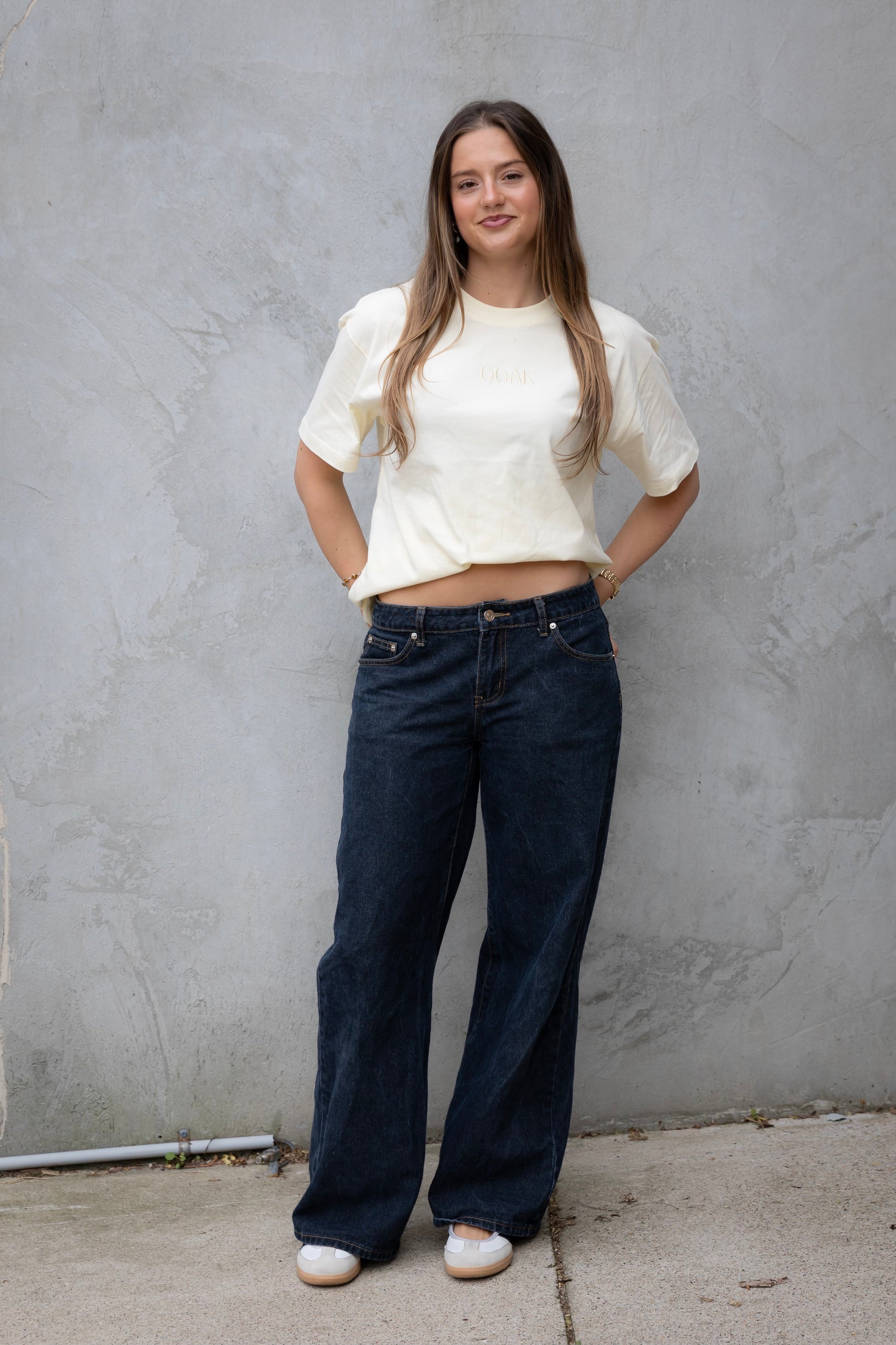 Woman wearing a white top and blue jeans standing against a gray wall.
