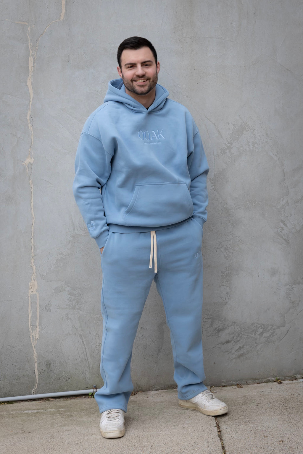 Man wearing a light blue hoodie and tracksuit standing against a gray wall.