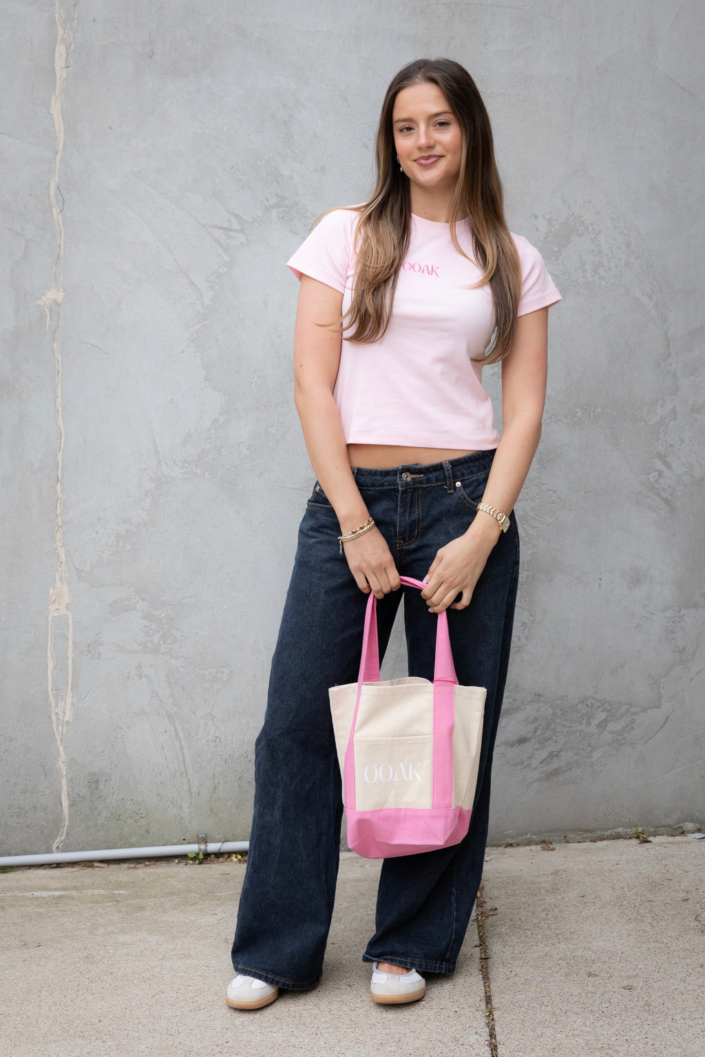 Woman holding a tote bag with a visible brand logo against a gray wall.