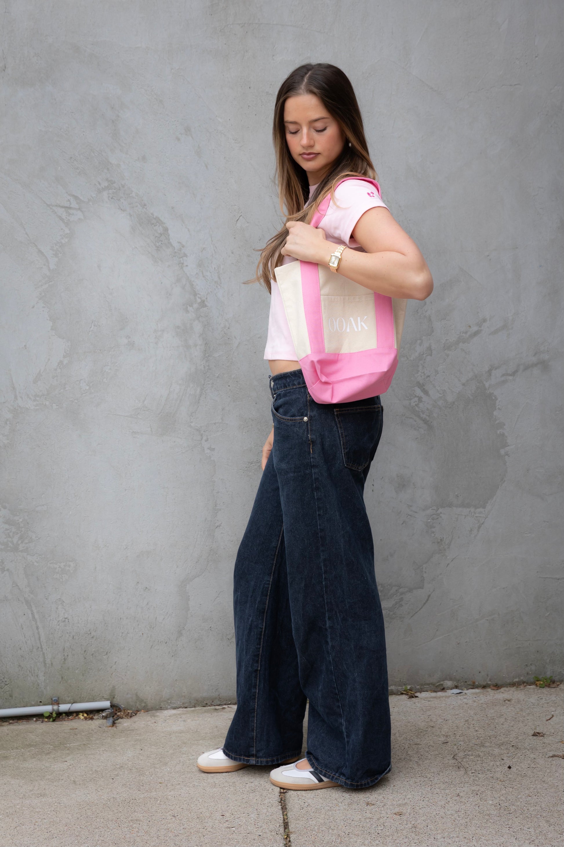 Woman wearing a light pink top with dark blue jeans against a gray wall.