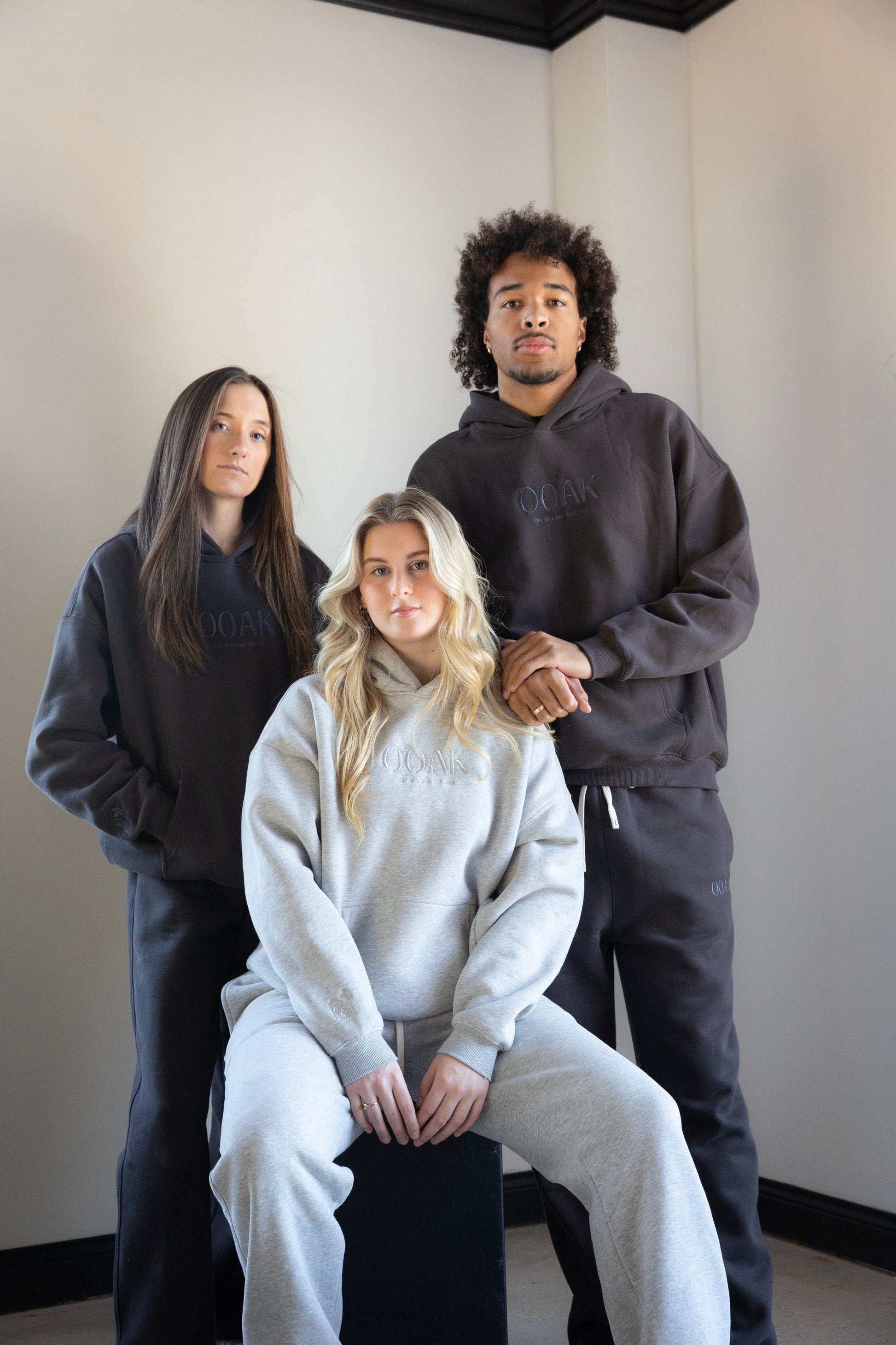 Three models wearing dark gray  & light gray hoodies with visible OOAK branding against a plain wall.