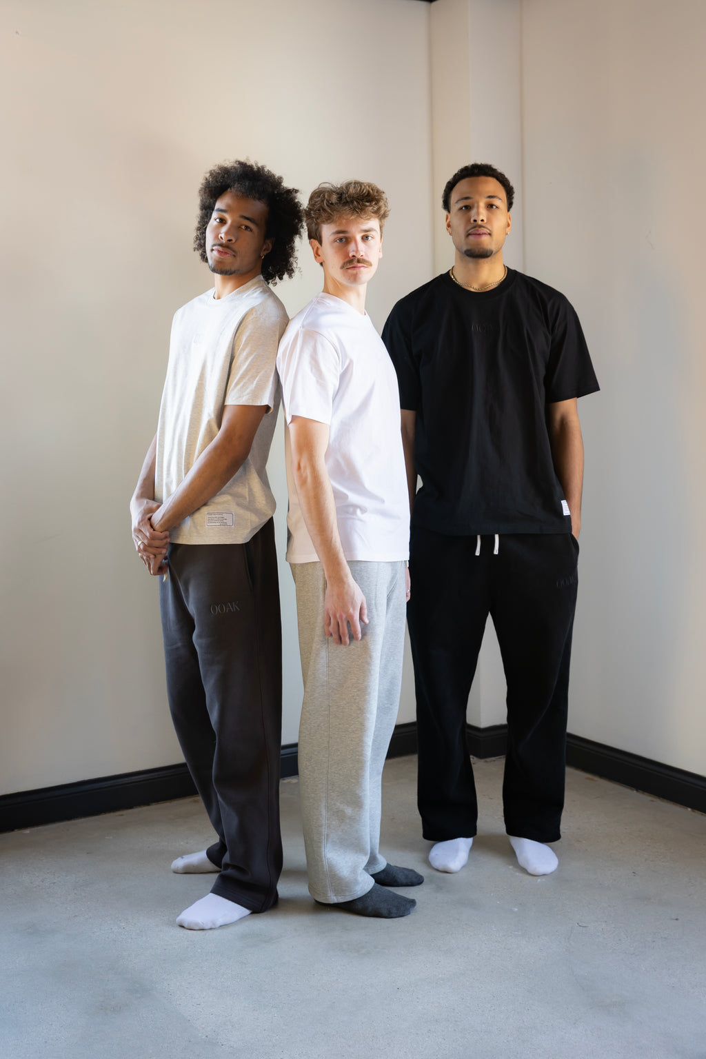 Three people standing against a plain wall wearing minimalistic outfits.