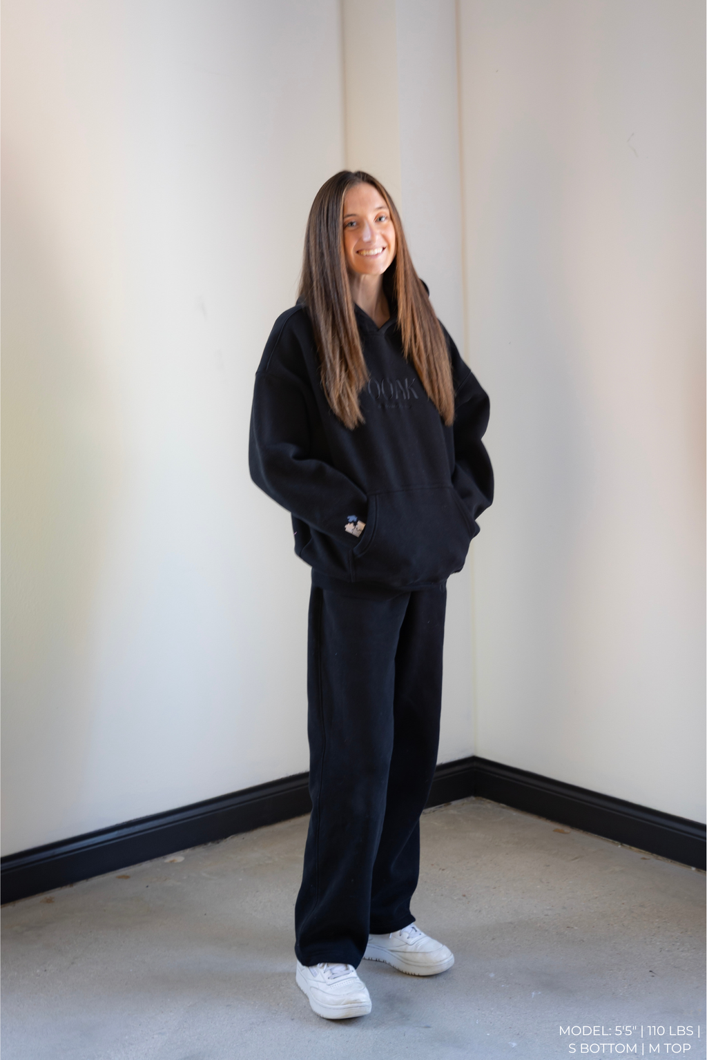 Person wearing a black outfit standing against a plain wall.