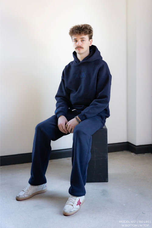 Model wearing a navy blue OOAK hoodie and pants sitting on a black box against a white wall.