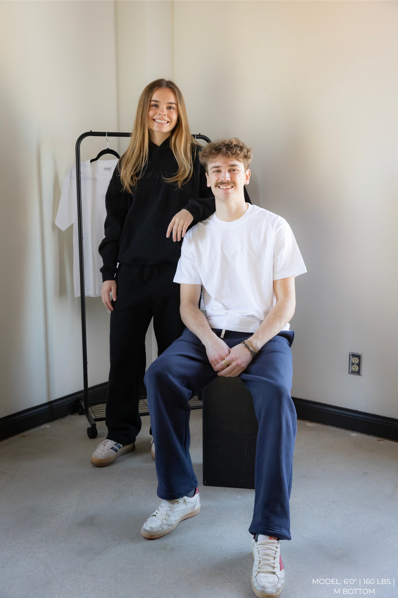 Two models in a minimalistic room with OOAK clothing on a rack.