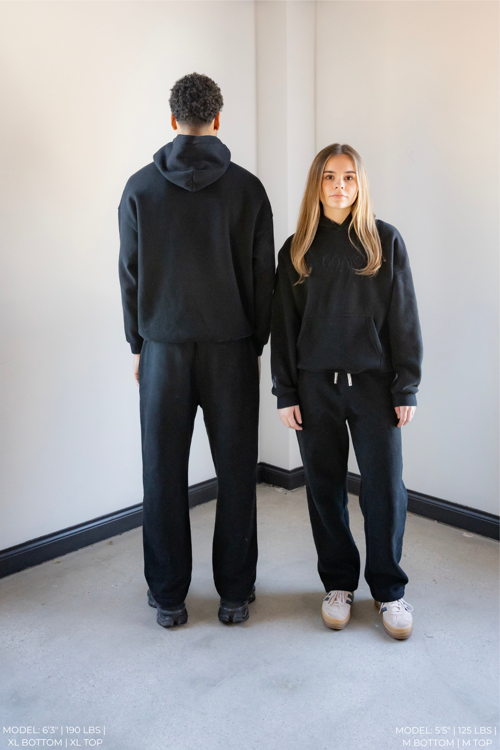 Two models wearing matching black OOAK sets standing back-to-back against a plain wall.