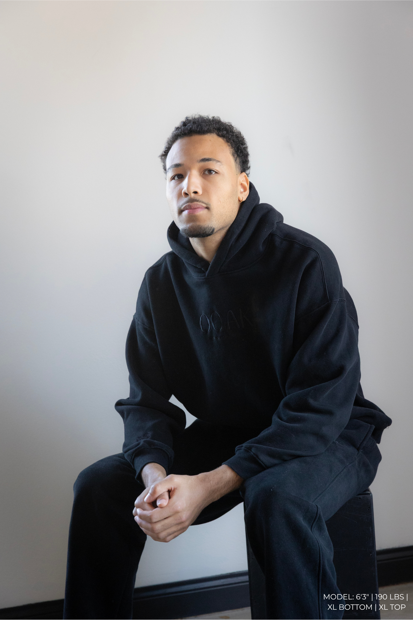 Man wearing a black hoodie and pants sitting against a plain background