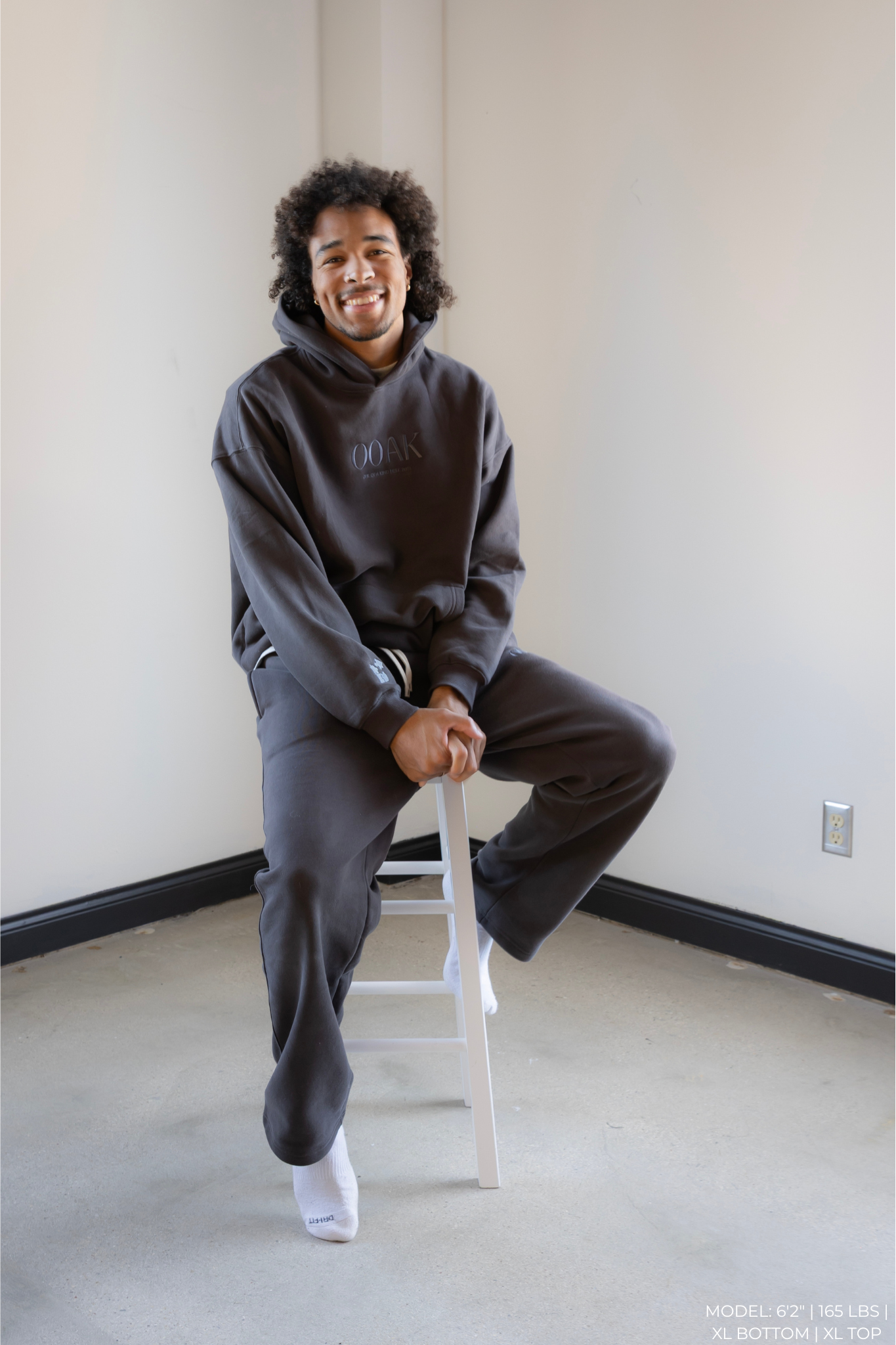 Person wearing a dark OOAK hoodie and pants sitting on a stool against a plain wall.